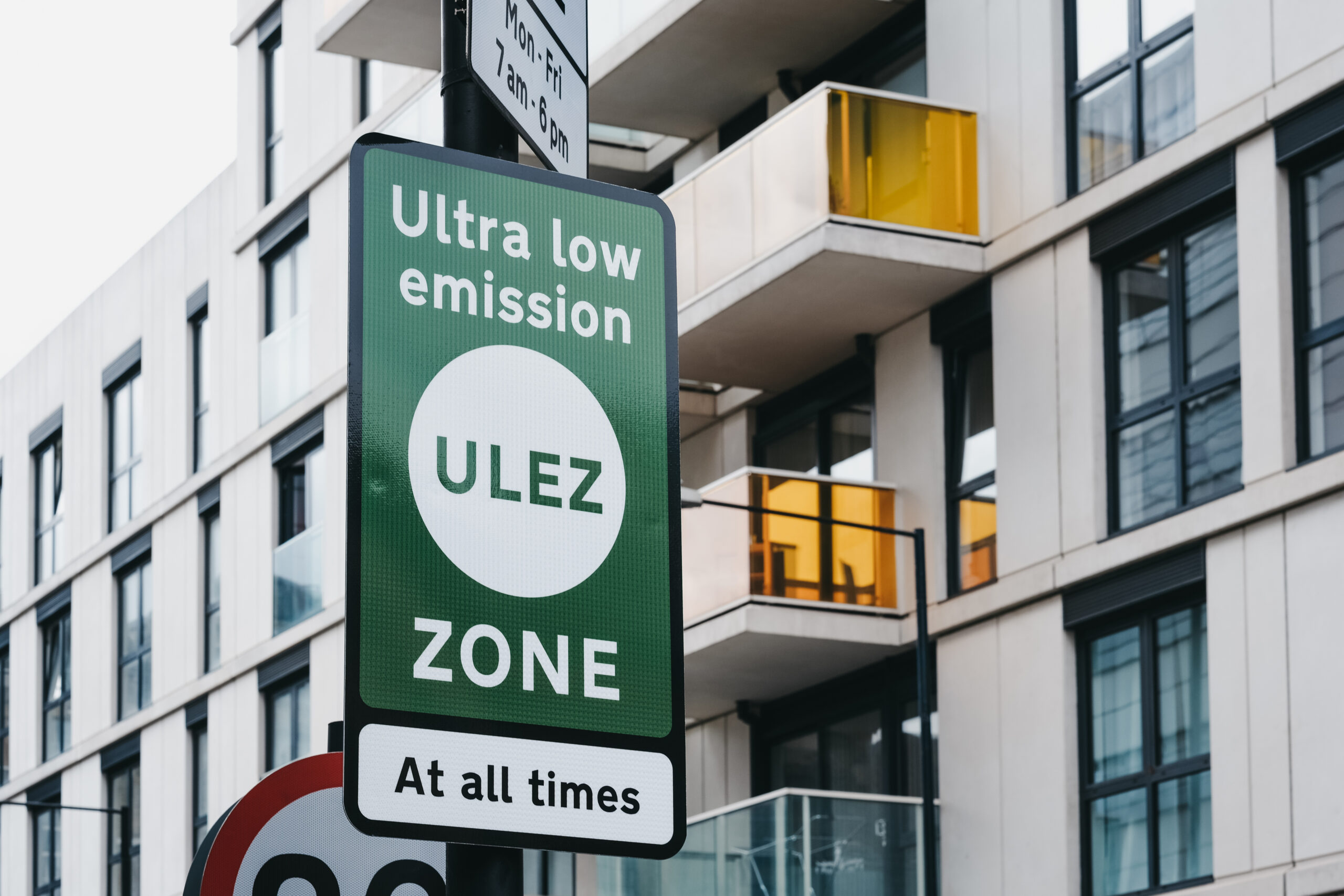 A green sign reads 'Ultra Low Emission ULEZ Zone At all times' in front of a modern building with glass balconies.
