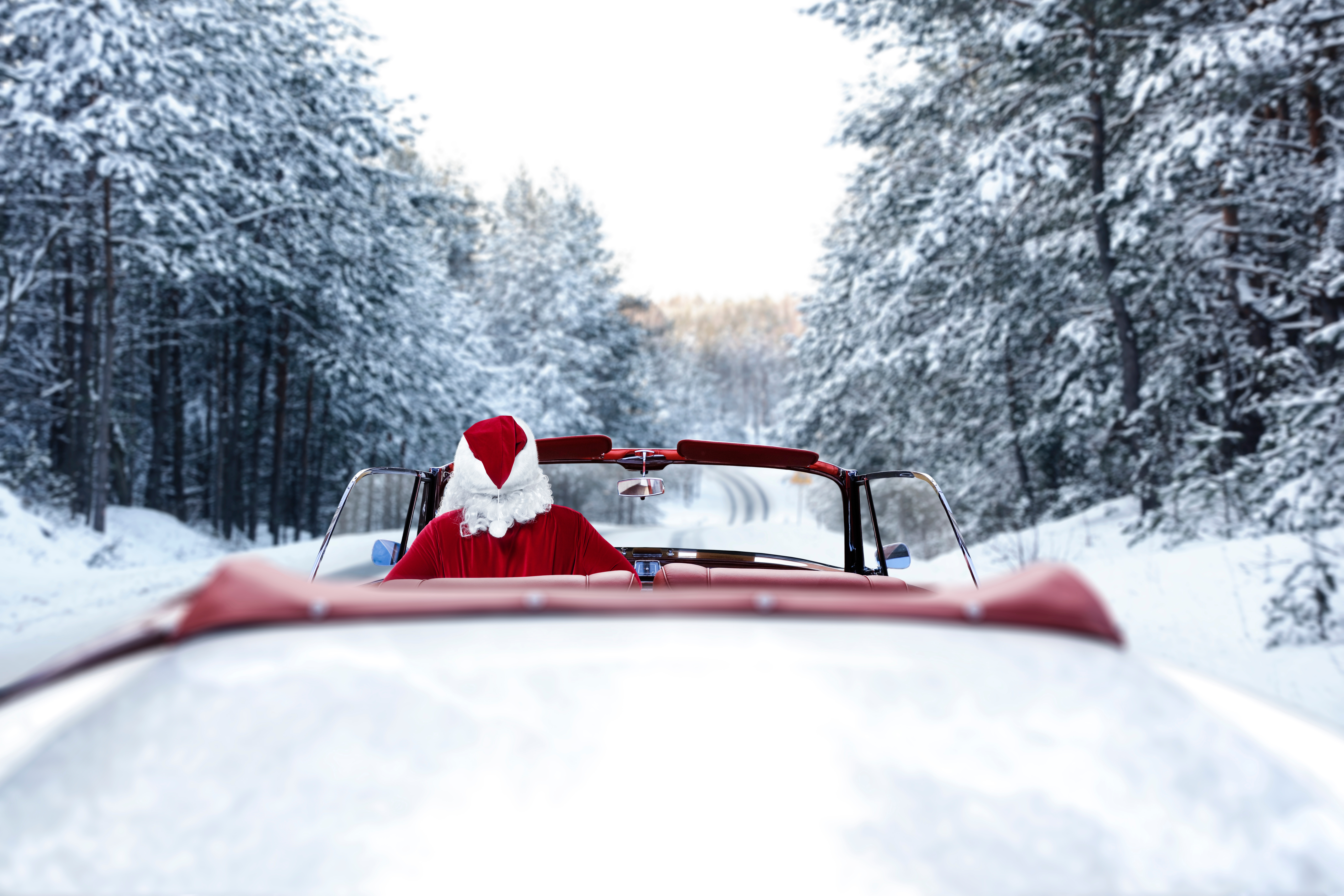 Santa Claus drives a red convertible through a snowy forest.