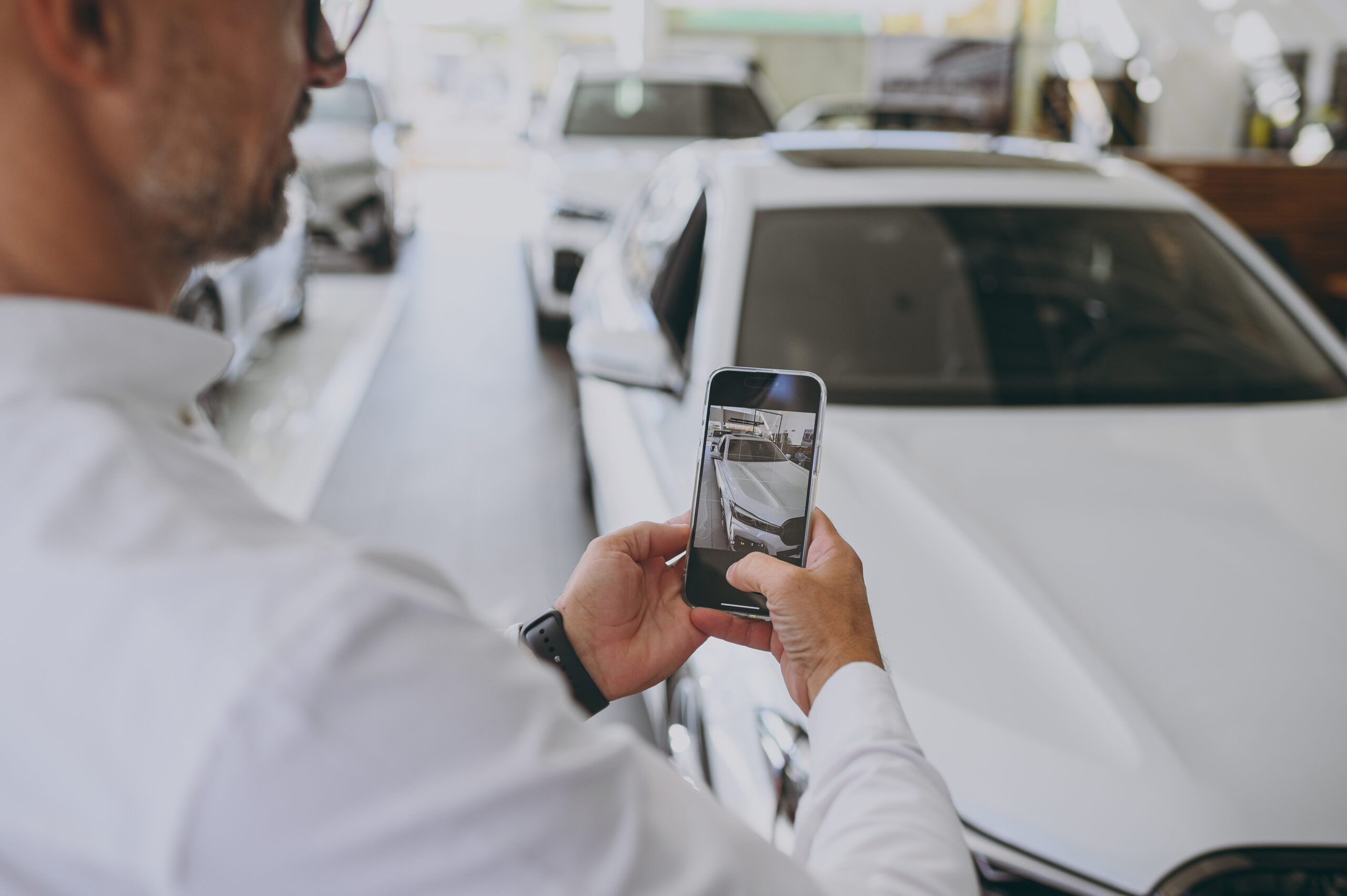 A person in a white shirt takes a photo of a white car with a smartphone in a showroom, conveying a focused and professional tone.