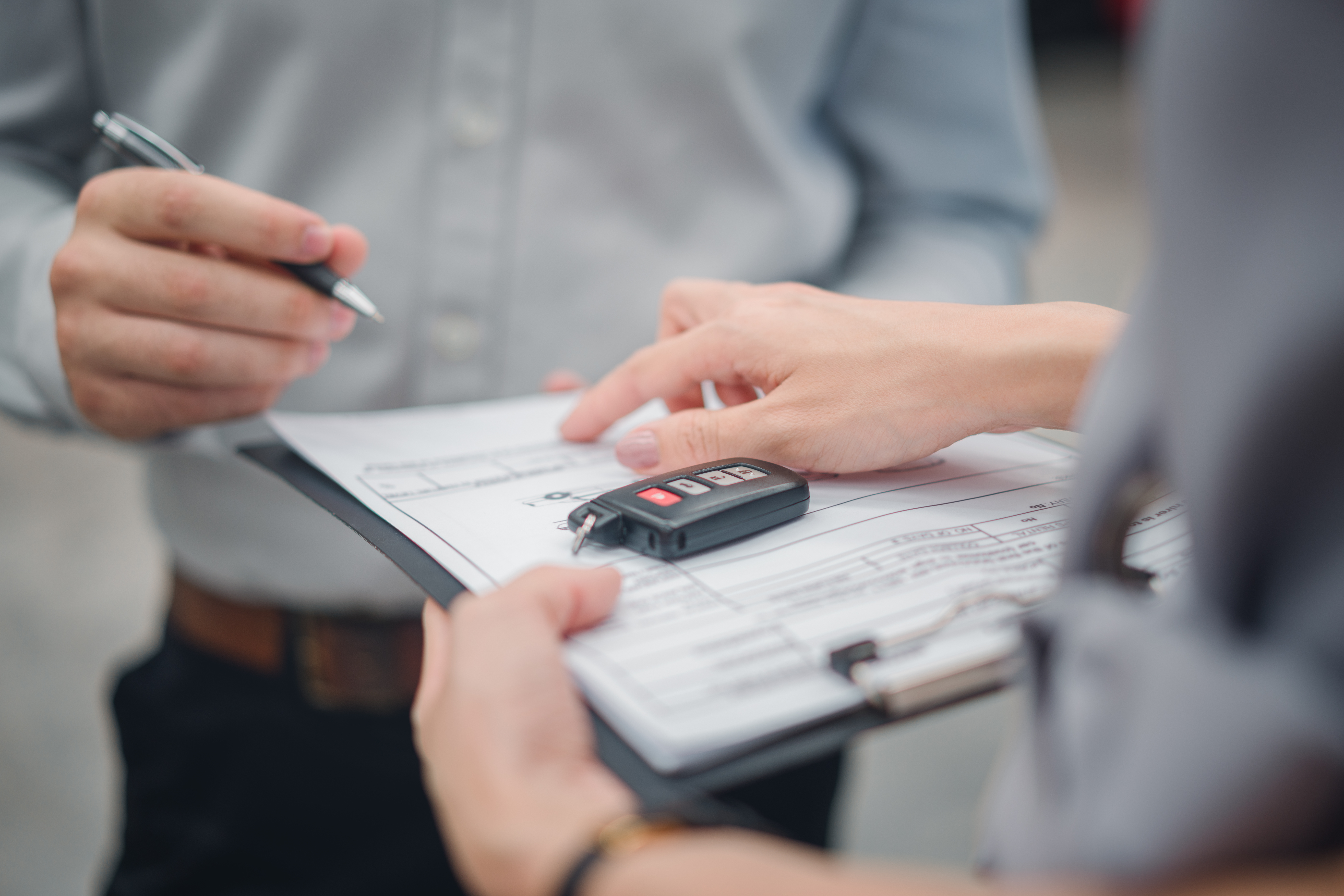 A person hands over car keys on a clipboard with documents to someone holding a pen.